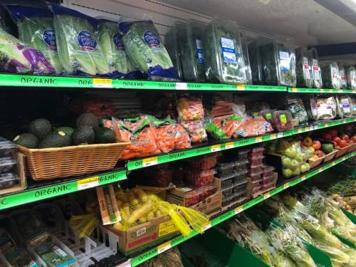 A grocery store shelf filled with various organic fruits and vegetables, including avocados, carrots, and apples.