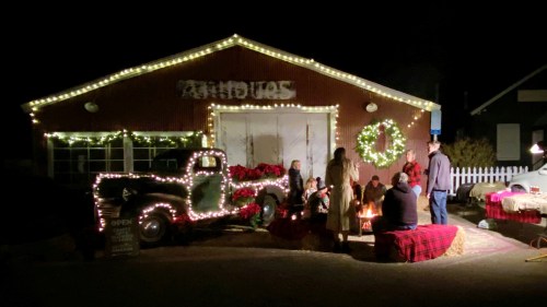 A festive scene at night with a decorated truck, people gathered around a fire, and holiday lights on a building.