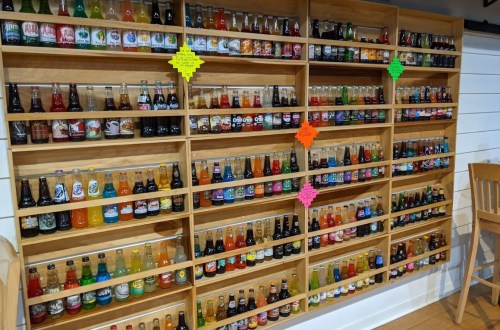 A wooden shelf displaying a colorful variety of soda bottles, organized by type and flavor, with bright labels.