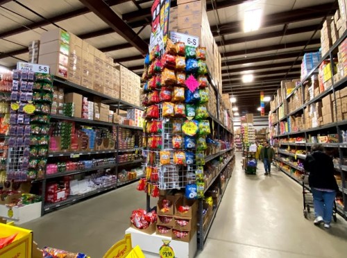 A wide aisle in a warehouse store filled with shelves of snacks and candy, with shoppers browsing.