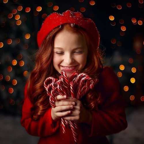 A girl in a red hat smiles joyfully while holding candy canes, surrounded by soft, glowing holiday lights.