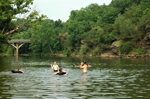 A group of people swimming and playing in a river surrounded by lush greenery and rocky cliffs. A bridge is visible in the background.