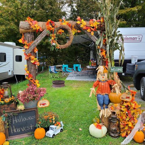 A festive Halloween harvest display with autumn leaves, a scarecrow, pumpkins, and a welcome sign in a campsite setting.