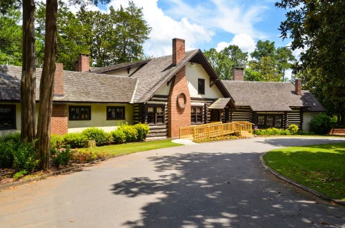 A charming log cabin-style building with a wooden ramp, surrounded by trees and a clear blue sky.