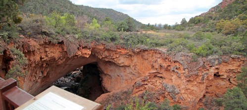 A scenic view of a natural rock arch over a lush canyon, surrounded by greenery and distant mountains.