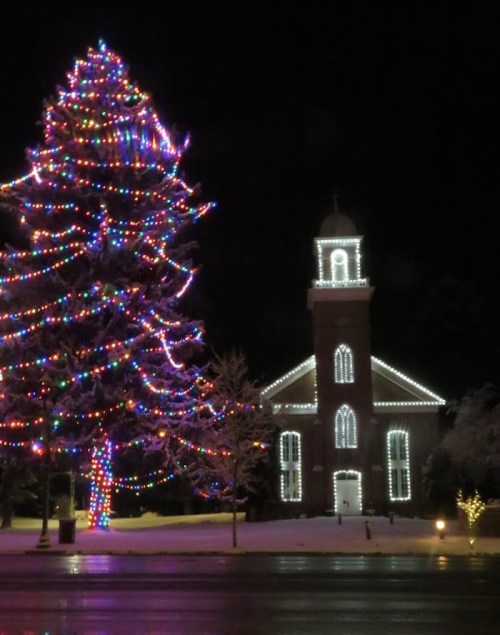 A brightly lit Christmas tree and a decorated church, both covered in colorful lights, set against a snowy backdrop.