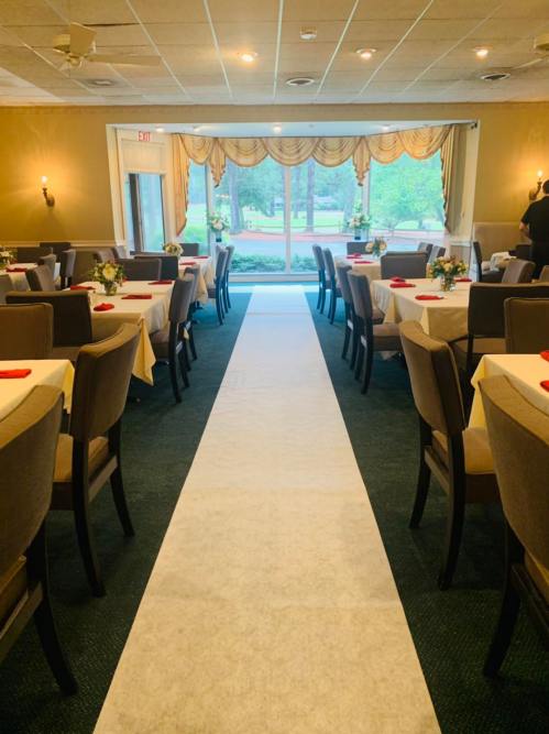 A banquet room with tables set for an event, featuring a white aisle runner leading to large windows.