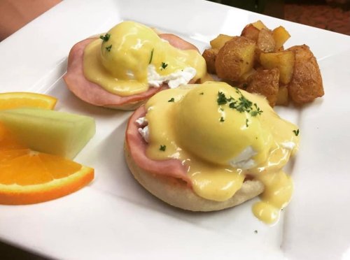 A plate of eggs Benedict with ham, hollandaise sauce, and a side of golden potatoes, garnished with fruit slices.