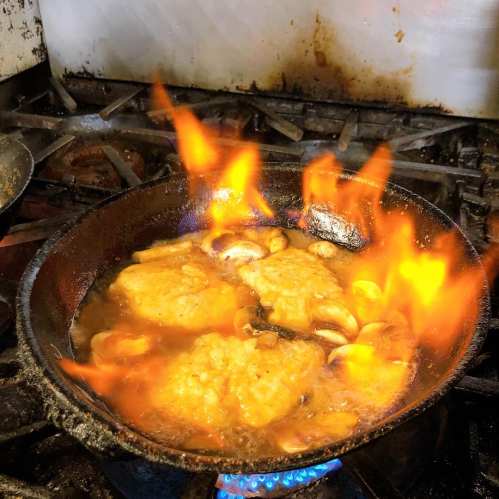 A skillet on a stove with chicken cooking, surrounded by flames and sizzling oil.
