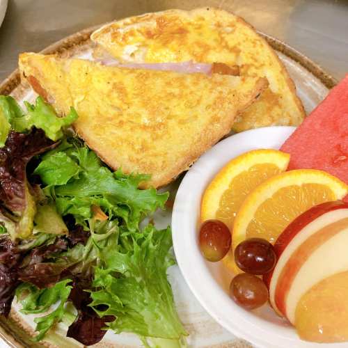 A plate with two slices of French toast, ham, mixed greens, and a bowl of fruit including oranges, grapes, and apple slices.