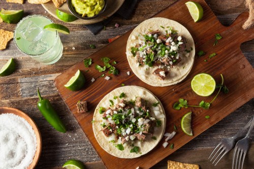 Two tacos on a wooden board, topped with meat, onions, cilantro, and cheese, surrounded by lime wedges and tortilla chips.