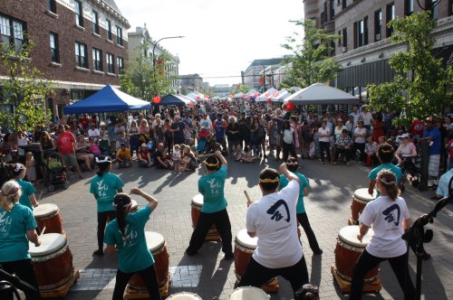 A lively street festival with a large crowd watching performers playing drums under colorful tents.