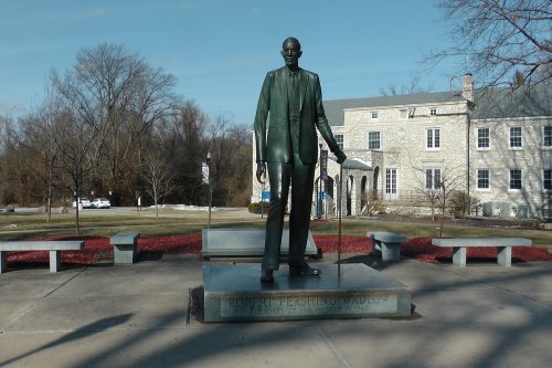 A bronze statue of a tall man in a suit, holding a cane, stands in a park with benches and trees in the background.