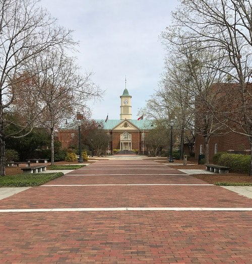 A brick pathway leads to a building with a clock tower, surrounded by trees and a cloudy sky.