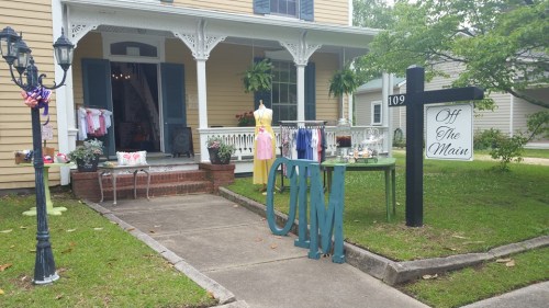 A charming storefront with a sign reading "Off The Main," featuring clothing displays and a welcoming porch.