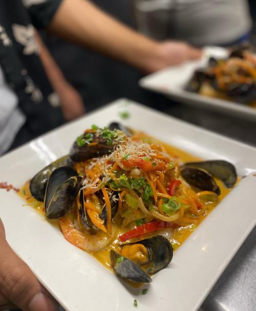 A plate of seafood pasta with shrimp, mussels, and colorful vegetables, garnished with herbs and cheese.