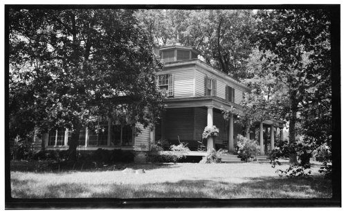 A historic house surrounded by trees, featuring a porch and large windows, captured in black and white.