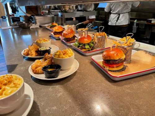 A variety of gourmet burgers with sides of fries, mac and cheese, and chicken, arranged on a countertop in a kitchen.