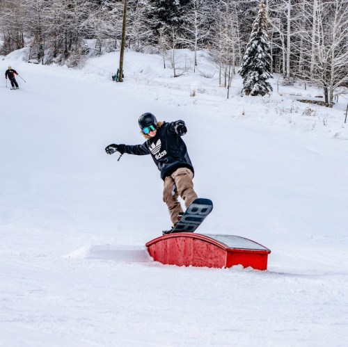 A snowboarder performs a trick on a red rail in a snowy landscape, with trees and another skier in the background.