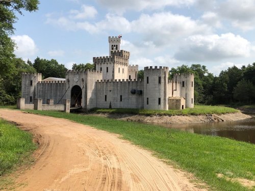 A large stone castle with towers and a flag, surrounded by greenery and a dirt path leading to a nearby water body.