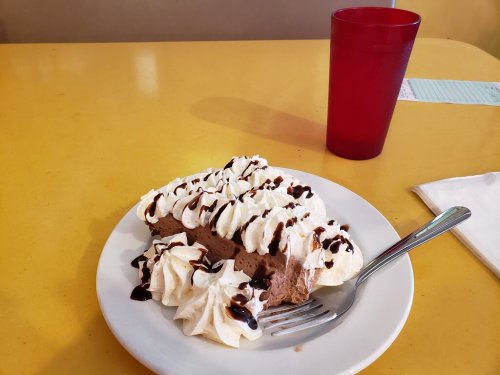 A slice of chocolate mousse cake topped with whipped cream and chocolate drizzle, next to a red cup on a yellow table.