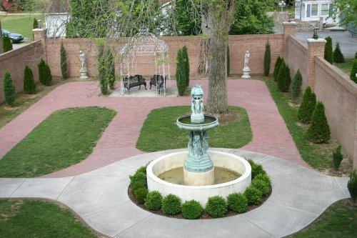 A landscaped garden featuring a central fountain, brick pathways, and decorative statues surrounded by greenery.