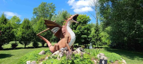 A large dragon sculpture made of bricks, surrounded by greenery and rocks, under a blue sky with fluffy clouds.