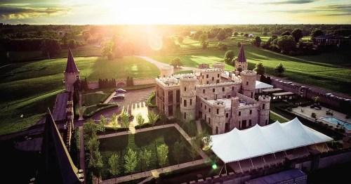 Aerial view of a castle with towers, surrounded by green fields and a large tent in the foreground, under a sunset sky.