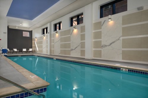 Indoor swimming pool with blue water, marble walls, and lounge chairs along the side, well-lit with large windows.