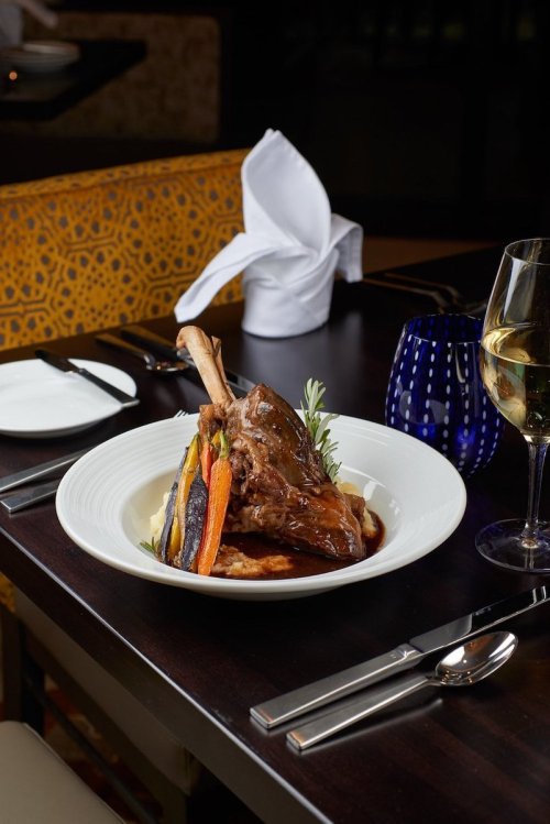 A plated dish featuring a lamb shank with vegetables, served on a white plate with a glass of white wine nearby.