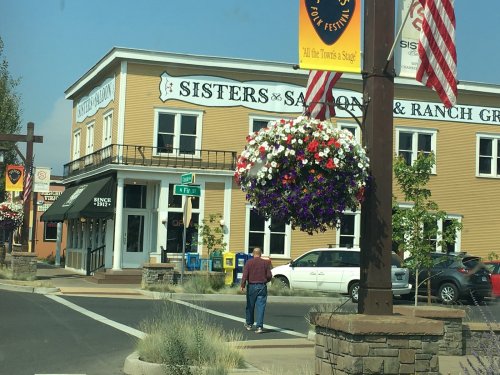 A man walks past a building with "Sisters Saloon & Ranch Grill" signage, surrounded by flowers and American flags.