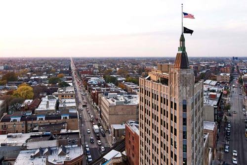 Aerial view of a city skyline featuring a tall building with an American flag and a long street lined with cars.