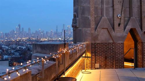 A rooftop view at dusk, featuring city skyline lights and decorative string lights along the edge.
