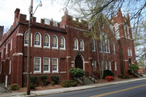 Historic red brick building with Gothic architecture, featuring arched windows and a steep roof, surrounded by greenery.