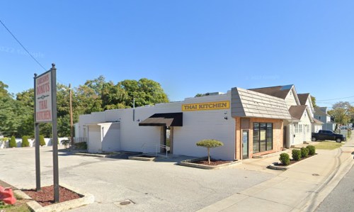 Exterior of a Thai restaurant with a sign reading "Thai Kitchen," surrounded by greenery and clear blue skies.