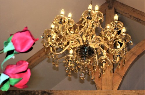 A sparkling chandelier with multiple lights hangs from a ceiling, with red roses partially visible in the foreground.