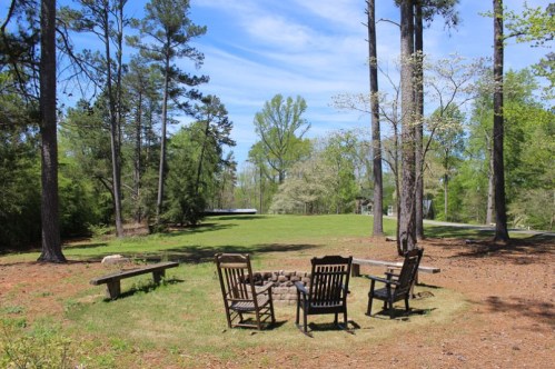 A serene outdoor space with wooden chairs around a fire pit, surrounded by trees and a grassy area under a blue sky.