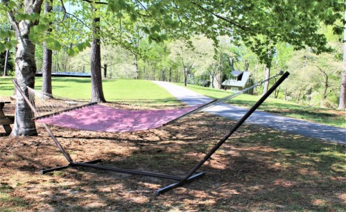 A pink hammock is set up between trees on a grassy area, with a path and a cabin in the background.