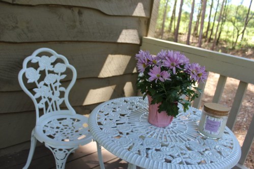 A white metal table and chair set on a porch, featuring a pink flower pot and a candle, surrounded by trees.