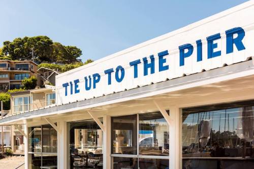 Sign on a building reads "TIE UP TO THE PIER," with a view of boats and hillside homes in the background.