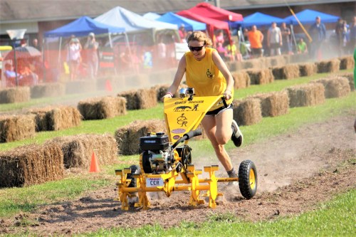 A person in a yellow tank top races a tiller on a dirt track, surrounded by spectators and hay bales.