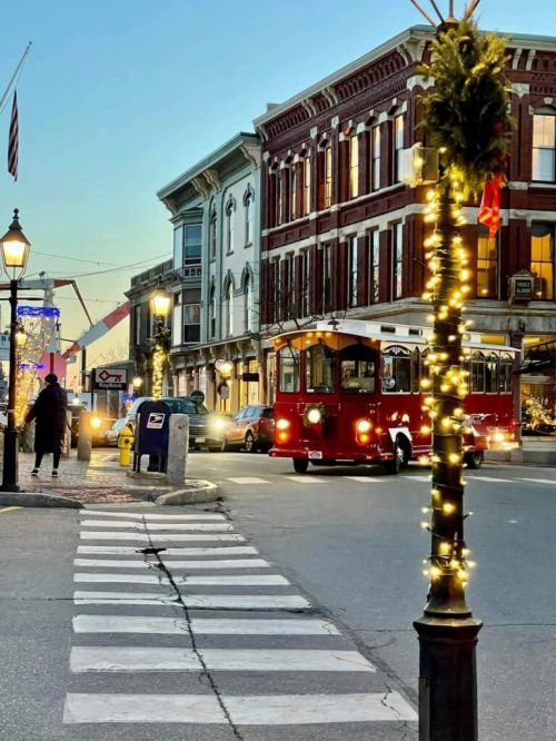 A festive street scene with a red trolley, decorated lampposts, and historic buildings at dusk.