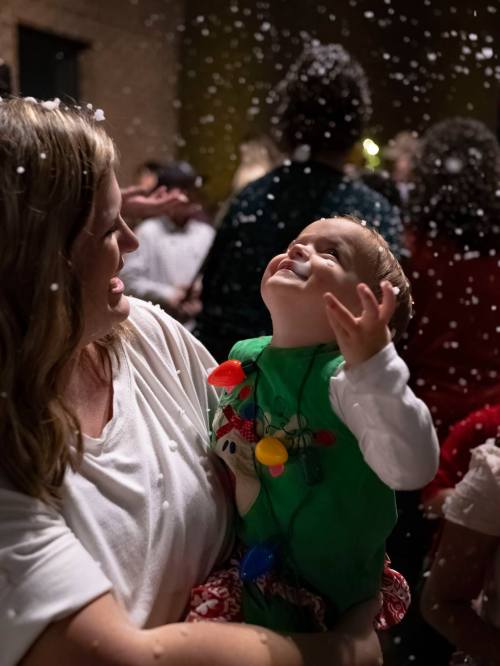 A woman holds a child in a festive outfit, both smiling as snow-like confetti falls around them.