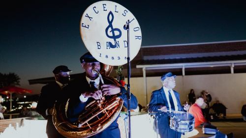 A brass band performs at night, featuring a tuba player and a drummer, with a large "Excelsior Band" sign.