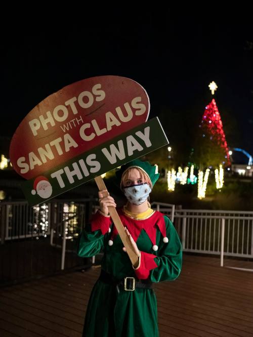 A person in an elf costume holds a sign directing to "Photos with Santa Claus" near a decorated Christmas tree.
