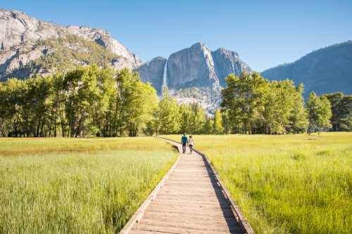 A couple walks along a wooden path through a green meadow, with mountains and a waterfall in the background.