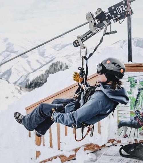 A person in winter gear rides a zip line over snowy mountains, with a scenic view in the background.