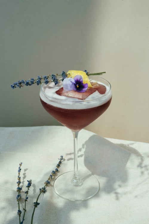 A cocktail garnished with a flower and lemon slice, served in a glass with lavender sprigs beside it.