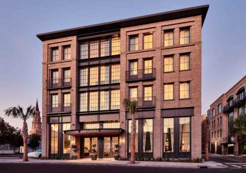 A modern brick hotel with large windows, palm trees, and a historic church in the background at sunset.