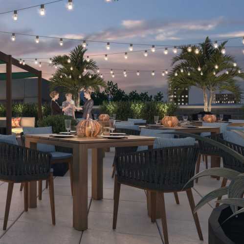 A rooftop dining area with string lights, tables, and people socializing among greenery and palm trees.